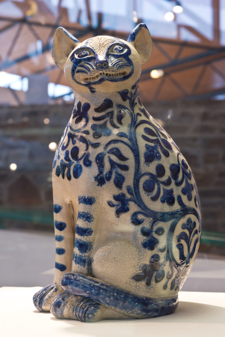 Keramikskulptur einer sitzenden Katze mit blauen floralen Mustern auf weißem Grund, aufgenommen in Innenraum mit unscharfem Hintergrund und Dachkonstruktion