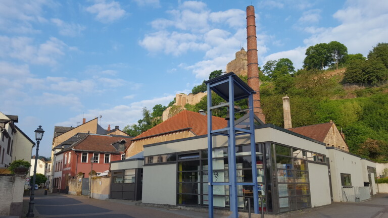 Stadtansicht mit modernen und traditionellen Gebäuden, einem blauen Glockenturm im Vordergrund, einem hohen roten Schornstein und einer Burgruine auf einem bewaldeten Hügel im Hintergrund unter einem blauen Himmel mit Wolken.