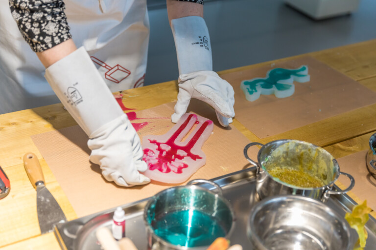 Person mit schützenden weißen Handschuhen, die eine Silikonform mit pinkfarbener Flüssigkeit auf einem Holzarbeitsplatz hält, daneben ein zweites Silikonobjekt mit blauer Flüssigkeit und mehrere Metallbehälter mit verschiedenen Flüssigkeiten und Werkzeugen.