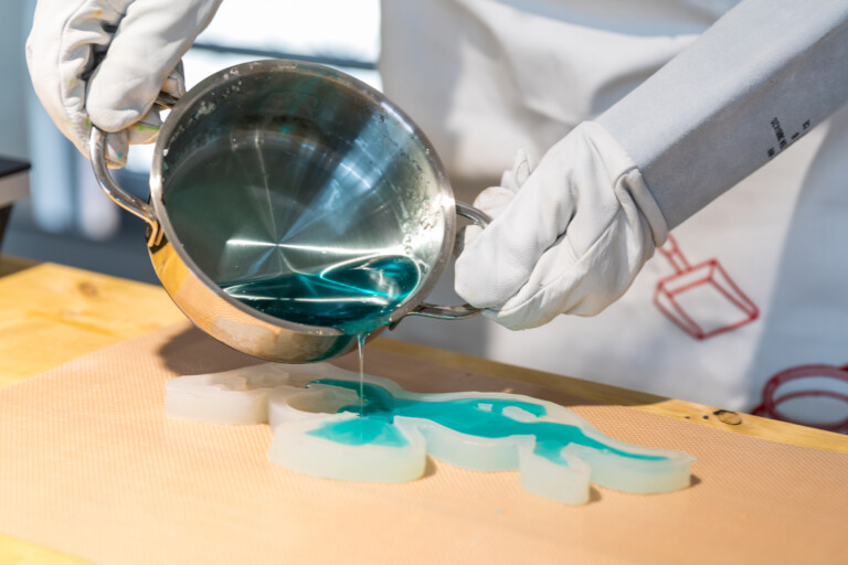 Person mit hitzebeständigen Handschuhen gießt blaue Flüssigkeit aus einem Metalltopf in eine Dinosaurus-förmige Silikonform auf einer Arbeitsfläche.