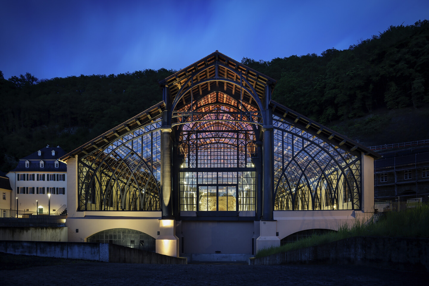 Beleuchtetes historisches Glas- und Stahlgebäude bei Nacht, umgeben von dunklen Hügeln und eingeschlossen von weiteren Gebäuden im Hintergrund.