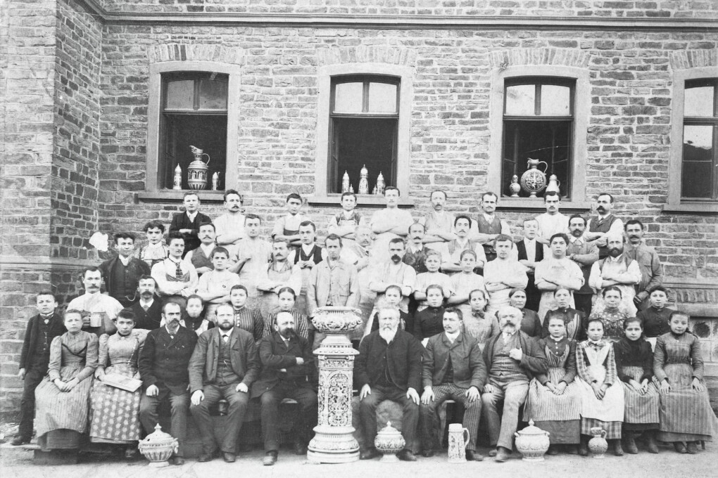 Schwarzweiß-Gruppenfoto vor einer Backsteinwand mit vielen Männern, Frauen und Kindern in historischer Kleidung, einige tragen Schürzen, davor sind dekorative Töpferwaren auf Sockeln platziert.