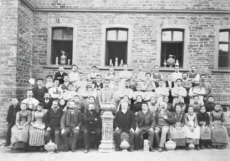 Schwarzweiß-Gruppenfoto vor einer Backsteinwand mit vielen Männern, Frauen und Kindern in historischer Kleidung, einige tragen Schürzen, davor sind dekorative Töpferwaren auf Sockeln platziert.