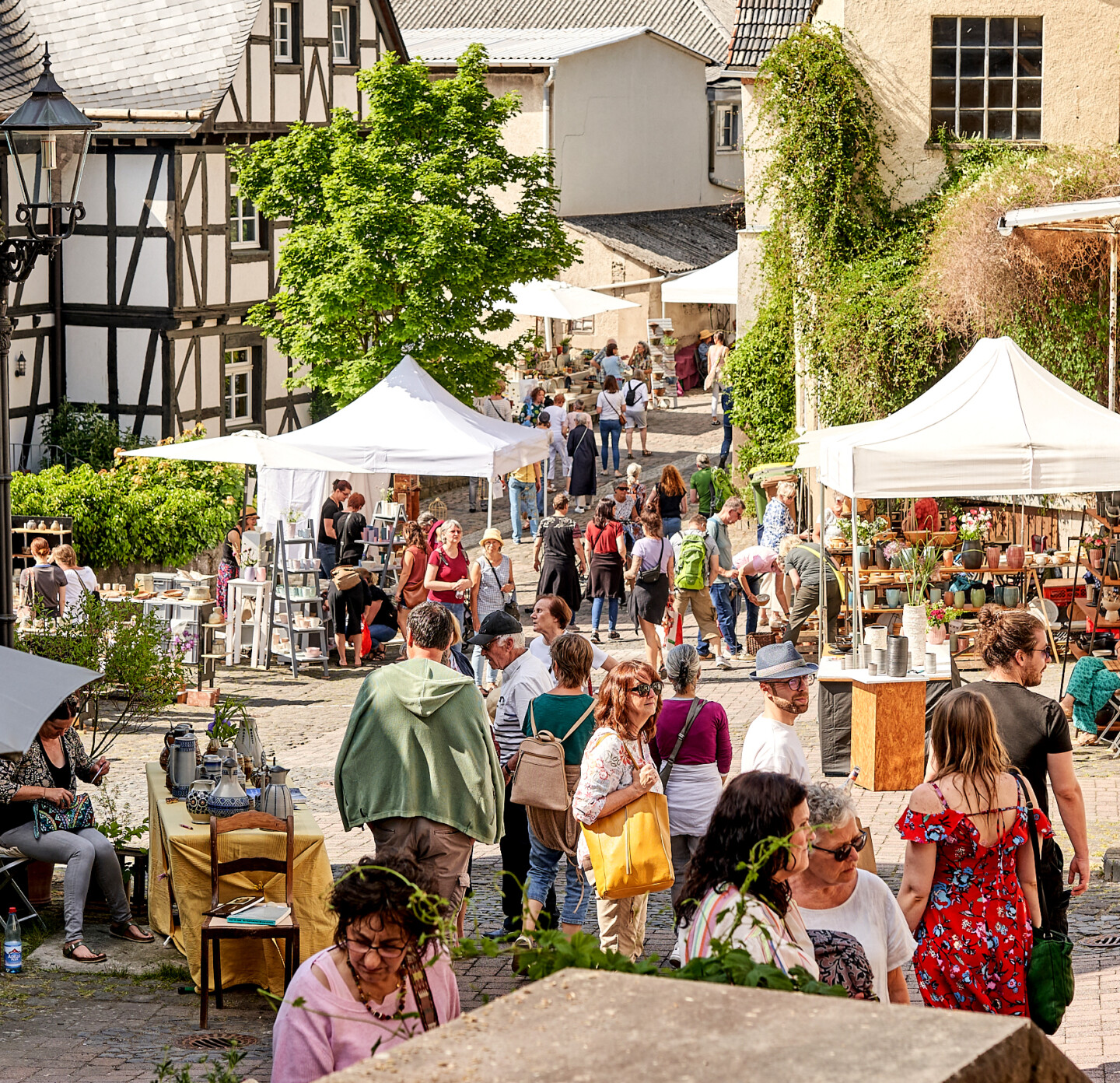 Belebter Straßenmarkt in einer historischen Stadt mit Fachwerkhäusern, weißen Verkaufszelten und Töpfereiwaren, umgeben von Besuchern in sommerlicher Kleidung bei sonnigem Wetter.