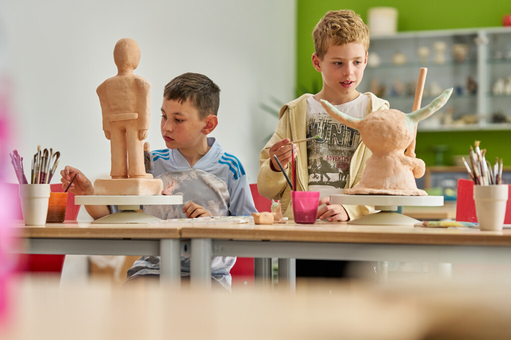 Zwei Jungs sitzen an einem Tisch und bemalen Terrakotta-Skulpturen in einem Klassenraum mit grüner Wand im Hintergrund, vor ihnen stehen Pinsel und bemalte Figuren auf Drehscheiben.
