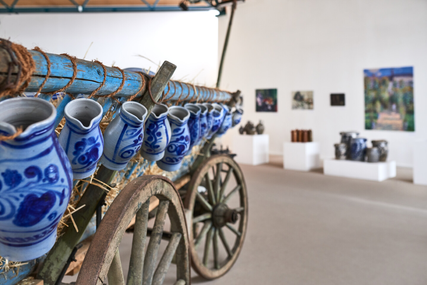 Nahaufnahme von blau-weiß bemalten Tongefäßen, die an einem alten Holzwagen mit großen Rädern befestigt sind, im Hintergrund unscharf eine Galerie mit Kunstwerken und weiteren Tongefäßen sichtbar.