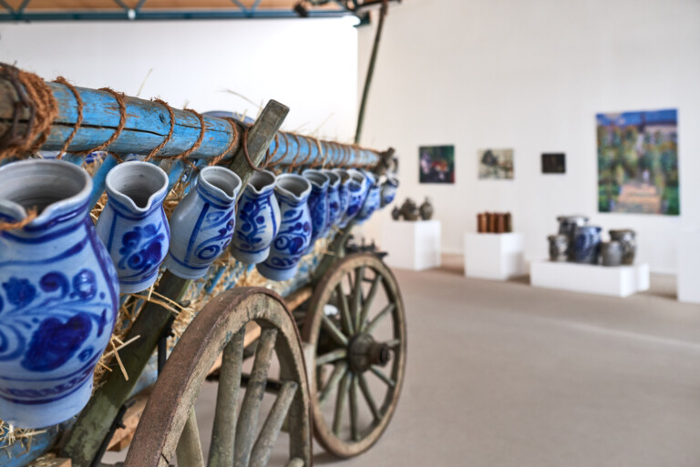 Nahaufnahme von blau-weiß bemalten Tongefäßen, die an einem alten Holzwagen mit großen Rädern befestigt sind, im Hintergrund unscharf eine Galerie mit Kunstwerken und weiteren Tongefäßen sichtbar.