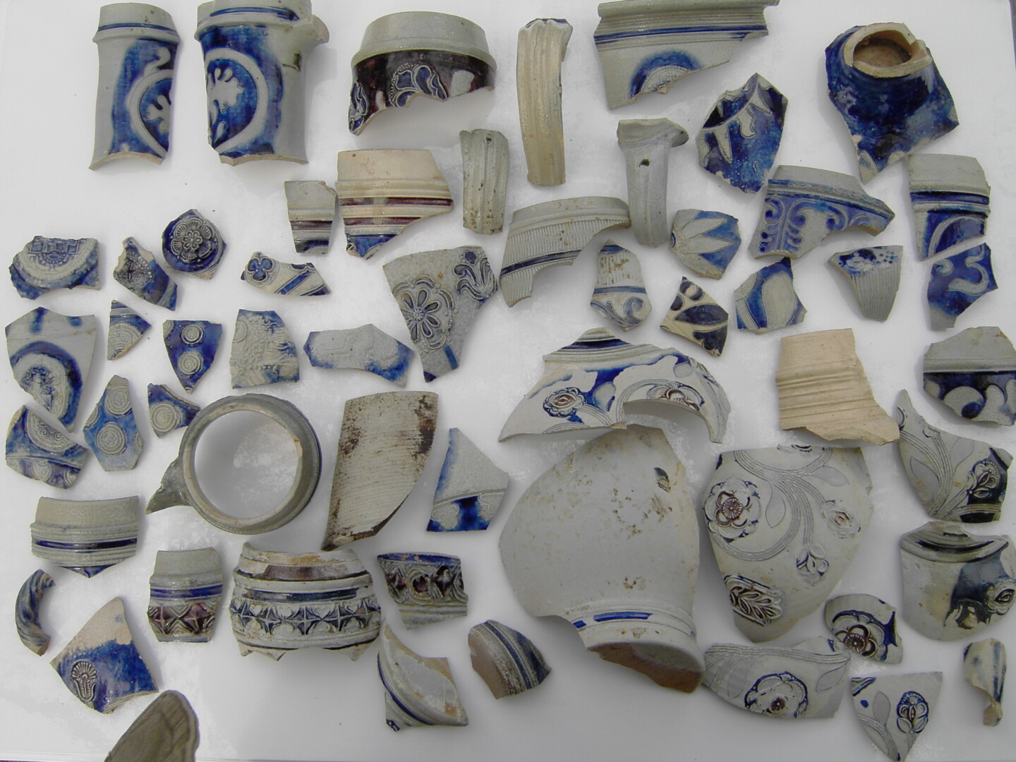 Mehrere zerbrochene Keramik- und Porzellanscherben mit blauen und weißen Verzierungen, verteilt auf einer weißen Oberfläche.