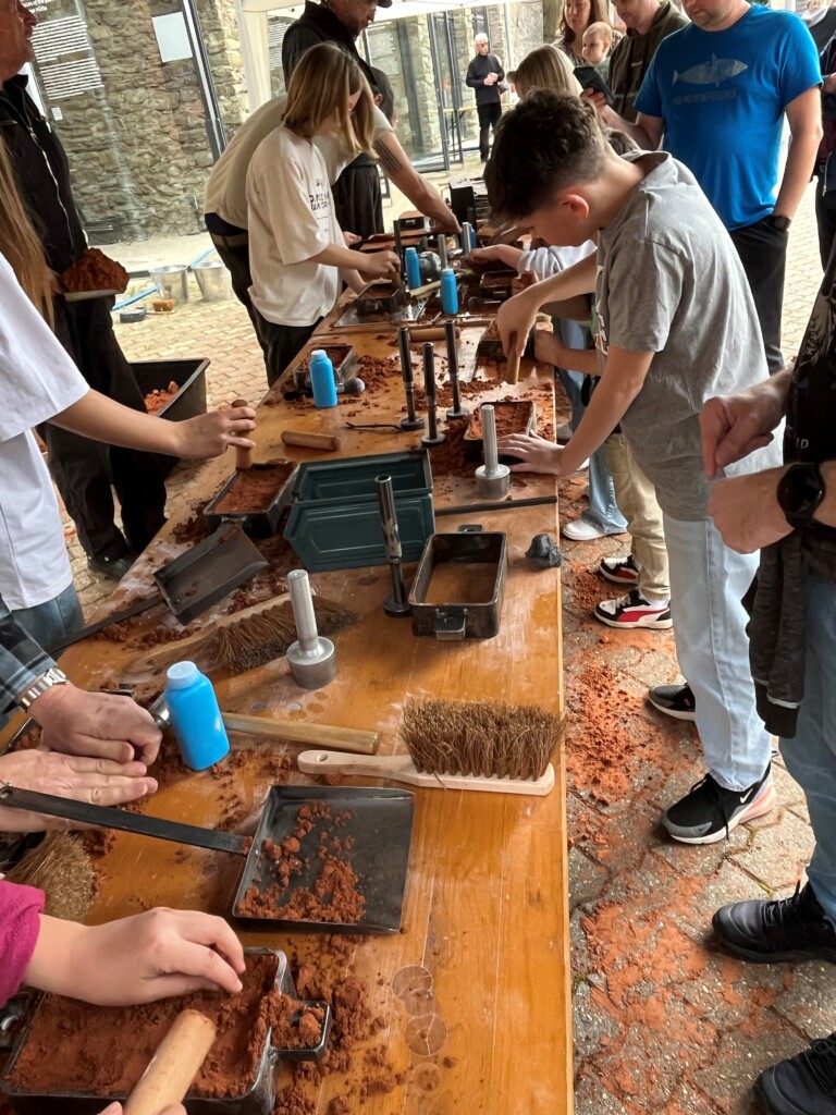 Menschen unterschiedlicher Altersgruppen arbeiten gemeinsam an einem langen Tisch mit Schokoladenformen und roten Materialien, beim Formen und Pressen von Schokolade in einem hellen Raum mit Steinwänden und Glasfenstern.