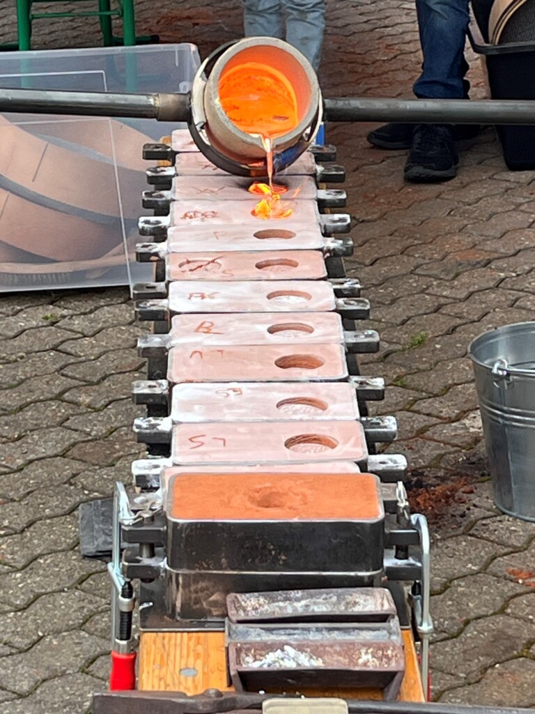 Ein Schmelztiegel gießt geschmolzenes Metall in eine Reihe von Gießformen auf einem gepflasterten Boden, im Hintergrund sind Teilansichten von Personen und ein Eimer zu sehen.