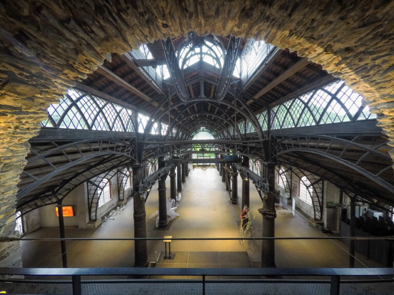 Innenansicht einer großen Halle mit kunstvoll gebogenen Metallbögen und Glasdach, seitlich Steinmauern mit Bogenfenstern, Blick von einem erhöhten Standpunkt durch einen steinernen Ausschnitt auf den leeren Hallenboden mit einigen Ausstellungsobjekten und Skulpturen.