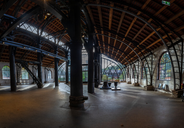 Großer, leerer Industriehalle mit Holzdecke und vielen schwarzen Metallstützen sowie großen Fensterfronten mit geschwungenen Eisenrahmen, durch die Tageslicht einfällt, rechte Seite mit zwei Stühlen vor der Wand.