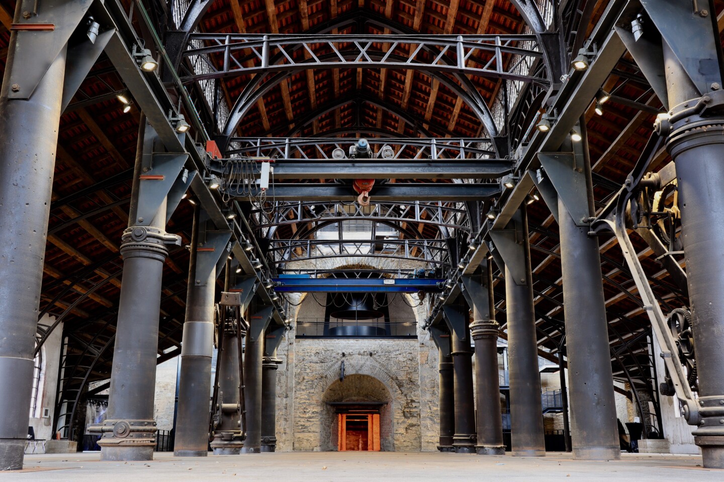 Industrielle Halle mit großen schwarzen Metallstützen, Stahlträgern und einem Gewölbedach mit Ziegeln, im Hintergrund ein Backsteinbogen mit rotem Lichtschein im Durchgang.