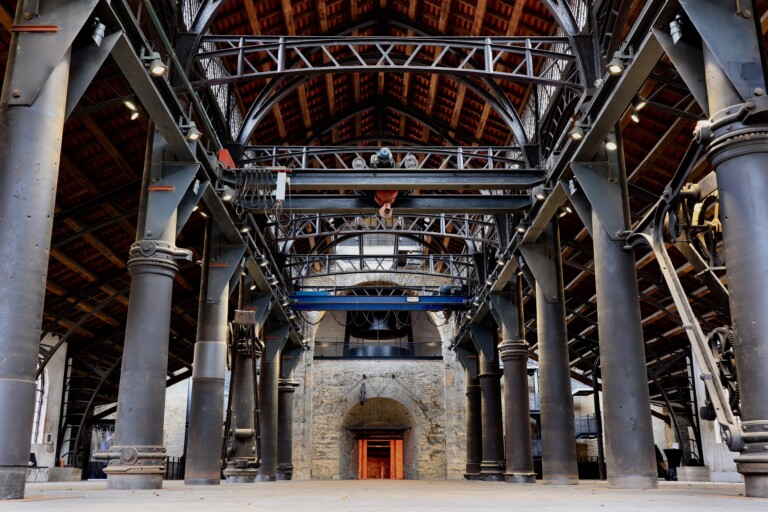 Industrielle Halle mit großen schwarzen Metallstützen, Stahlträgern und einem Gewölbedach mit Ziegeln, im Hintergrund ein Backsteinbogen mit rotem Lichtschein im Durchgang.