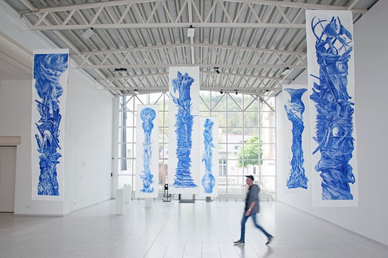 Große, vertikal hängende Stoffbanner mit blauen abstrakten Kunstmustern in einer hellen Galerie mit hohen Fenstern und sichtbarer Stahlträgerdecke, eine Person in Bewegung im Vordergrund