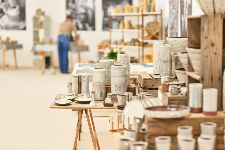 Näheaufnahme einer Ausstellung oder eines Ladens mit vielfältigen handgefertigten Keramikvasen, Schalen und Tassen auf Holztischen und Regalen, im Hintergrund unscharf eine Person und weitere Ausstellungsflächen