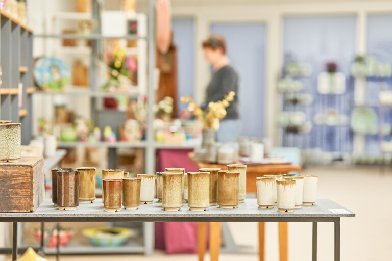 Verschiedene keramische Becher in Erdtönen auf einem Tisch in einem hellen Raum mit Regalen und einer unscharfen Person im Hintergrund