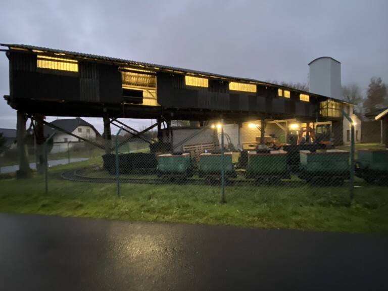 Dämmerungsaufnahme eines alten, verlängerten Holzgebäudes auf Stelzen mit beleuchteten Fenstern, davor ein Zaun und mehrere grüne Loren auf einer kleinen Gleistrasse, im Hintergrund Häuser und ein grauer Himmel
