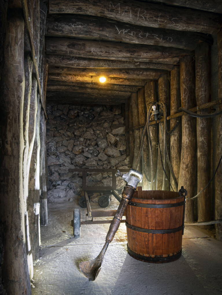 Innenraum eines Schachts mit steinernem Boden, einer hölzernen Tonne und einem alten, rostigen Bohrhammer, der schräg gegen die Tonne lehnt, mit einer Zugkarre im Hintergrund
