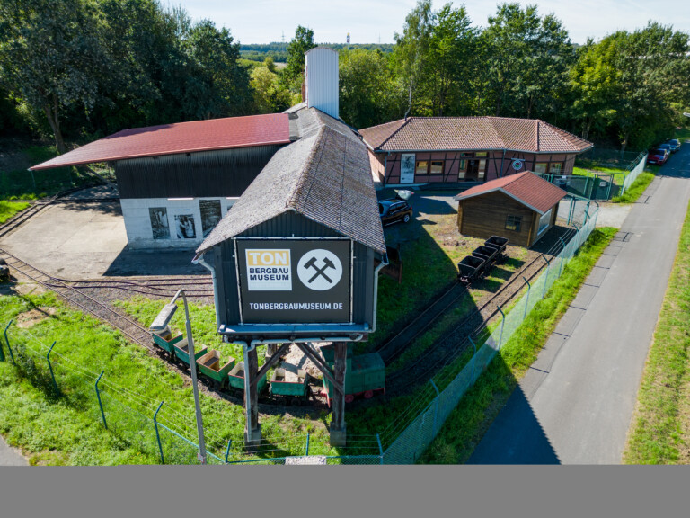 Außenansicht des Tonbergbau Museums mit historischen Loren auf einem kleinen Schienennetz, flankiert von einem Gebäude mit einem großen Schild und einem Fachwerkhaus, umgeben von Bäumen und grünem Gras an einem sonnigen Tag.