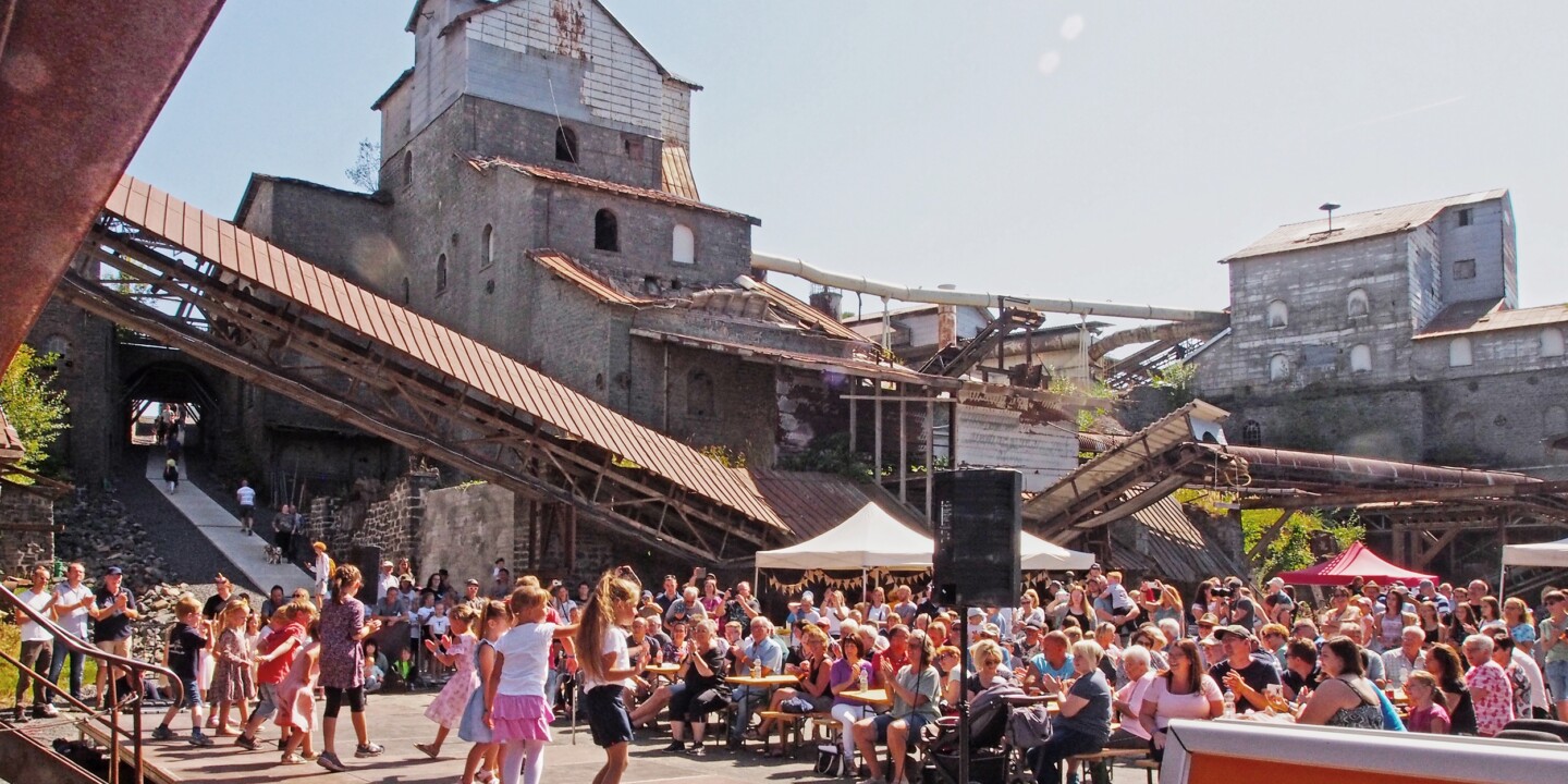 Große Menschenmenge bei sonnigem Fest vor altem Industriegebäude mit Stahlträgern und Förderbändern, Kinder tanzen vorne.
