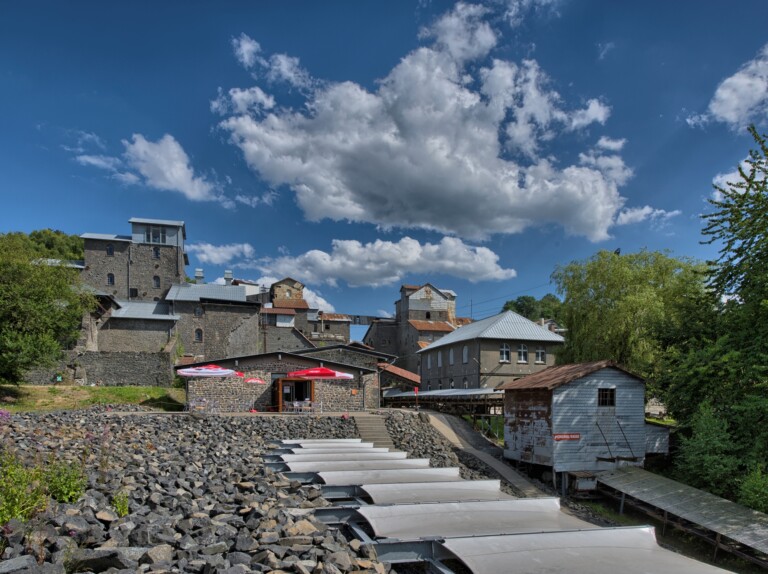 Blick auf ein historisches Industriegebäudeensemble aus Stein mit Sonnenschirmen und stahlverstärktem Gehweg, darunter ein klarer blauer Himmel mit großen Wolken.