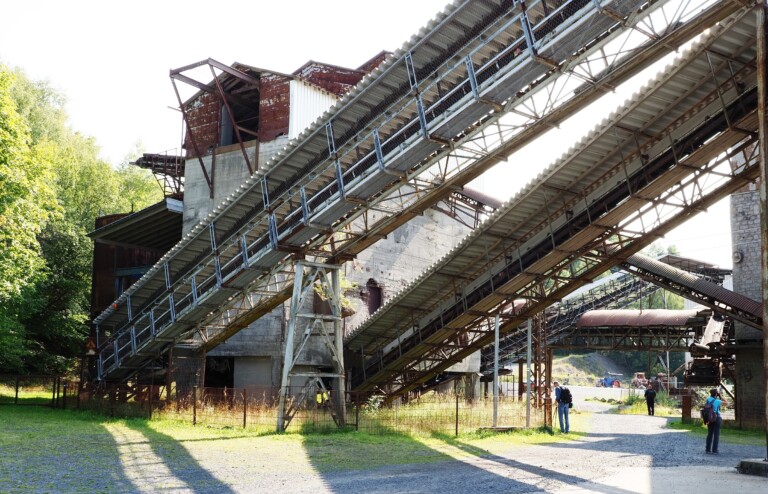 Industrieruine mit rostigen, schrägen Förderbändern und drei sichtbaren Personen im Außenbereich bei Sonnenschein.