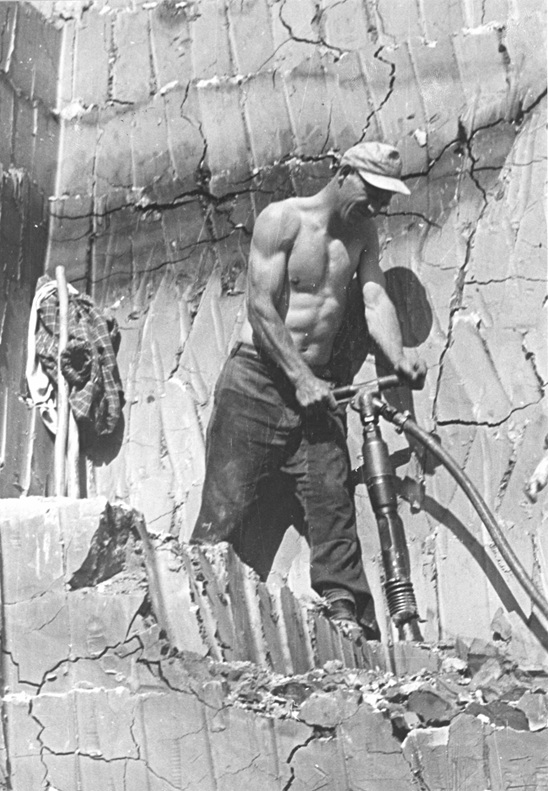 Oberkörperfreier Bauarbeiter bedient Presslufthammer an einer Felswand mit Werkzeug und Kleidung im Hintergrund.