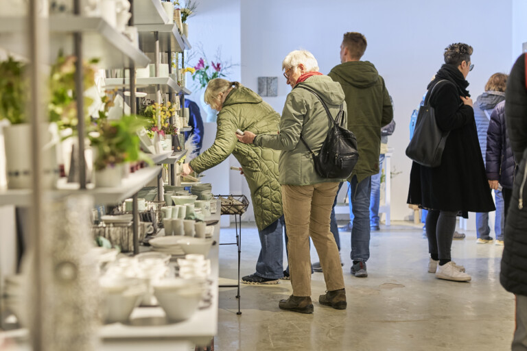 Menschen in einer Galerie oder Ladenumgebung betrachten Geschirr und Dekorationen auf weißen Regalen.