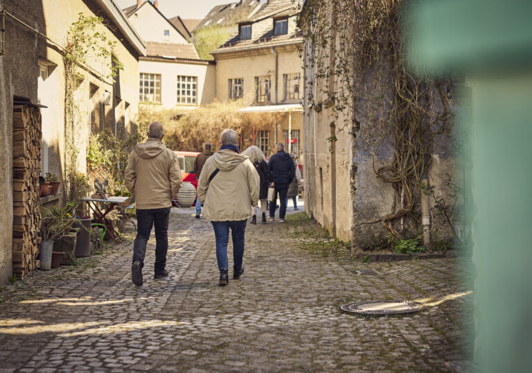 Gruppe von Menschen in Jacken beim Spaziergang auf gepflastertem Weg zwischen alten Gebäuden in sonnigem Innenhof.