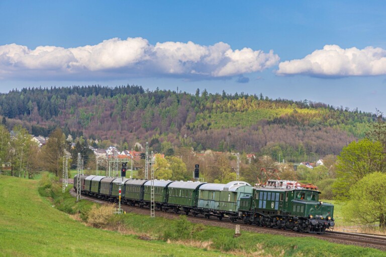 Grüner historischer Personenzug mit E-Lok auf Gleis vor Wald und blauem Himmel mit Wolken in ländlicher Umgebung.